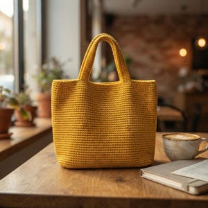 May include: A mustard yellow tote bag crafted with a dense, uniform crochet stitch pattern. The bag features two integrated handles at the top and a structured, rectangular base, showcasing a consistent texture throughout the entire exterior surface.