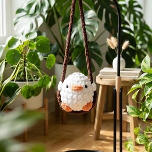 May include: A handmade white crochet bird with orange feet and beak, suspended in a brown crochet swing. The bird is hanging in front of lush green plants, creating a whimsical and cozy scene.