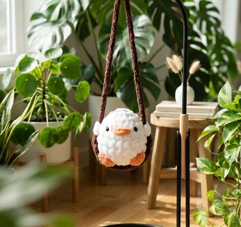 May include: A handmade white crochet bird with orange feet and beak, suspended in a brown crochet swing. The bird is hanging in front of lush green plants, creating a whimsical and cozy scene.