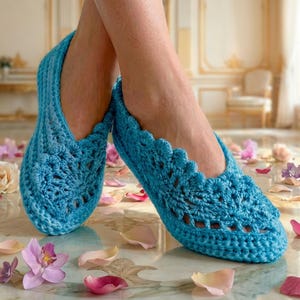 Puede incluir: Un par de zapatillas de ganchillo azul claro con un borde festoneado y un diseño calado. Las zapatillas están hechas a mano y parecen suaves y cómodas. La imagen está sobre una superficie de mármol con pétalos de flores esparcidos.