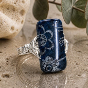 Pode incluir: Um anel retangular azul-escuro com um padrão floral branco na superfície. A pedra é fixada por uma banda de fio prateado que se enrola à volta dos lados da peça retangular, criando uma cravação de metal detalhada.