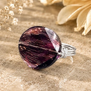 Pode incluir: Um anel com um cristal grande, redondo e facetado em roxo escuro com inclusões em tons de cobre metálico. A pedra é fixada por uma banda de fio prateado que se enrola em torno da base do cristal para mantê-lo firmemente no lugar.
