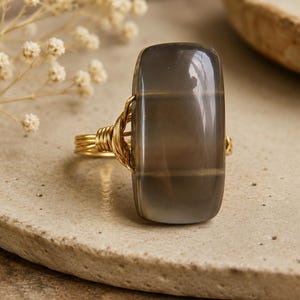 May include: A gold wire-wrapped ring featuring a large, rectangular, gray gemstone with horizontal bands of color. The ring's band is made of multiple strands of gold wire, creating a textured appearance. The gemstone has a polished, glossy surface.