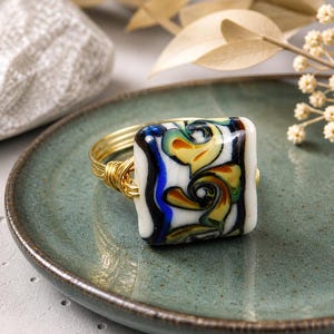 May include: A square ring with a gold wire band and a colorful, patterned glass top. The glass features swirls of yellow, orange, blue, and green against a white background. The ring is displayed on a teal plate.