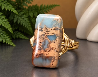 May include: A ring featuring a rectangular, polished gemstone with a marbled pattern of light blue and copper-brown tones. The stone is secured by a gold-colored wire-wrapped band that coils around the sides of the stone to hold it in place.