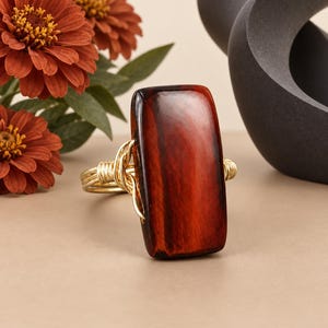 May include: A ring featuring a large, rectangular, polished red tiger's eye gemstone set in a gold-toned wire-wrapped band. The stone displays deep reddish-brown tones with a chatoyant luster, secured by intricate gold wire coils.