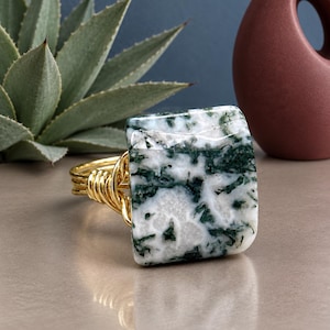 May include: A ring featuring a square-shaped moss agate stone with white and dark green inclusions. The stone is set on a band constructed from multiple strands of gold-colored wire that are wrapped around the base of the square gemstone setting.