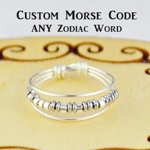 May include: A silver ring with three bands and silver beads. The ring is on a wooden surface. The text "CUSTOM MORSE CODE ANY ZODIAC WORD" is above the ring.