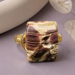 May include: A ring featuring a large, rectangular, polished stone with marbled patterns in shades of cream, olive green, and deep maroon. The stone is secured by multiple strands of bright gold-toned wire that wrap around the sides to form the band.