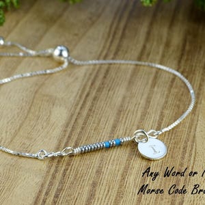 May include: A silver adjustable bracelet with a small silver charm that says "I" and a blue and silver beaded section. The bracelet is on a wooden surface. Any Word or Name Morse Code Bracelet.
