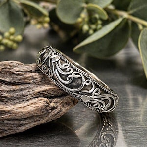 Peut inclure: Une bague en argent ornée d'un motif tourbillonnant complexe. La bague repose sur un morceau de bois flotté, avec un feuillage vert en arrière-plan. Le design de la bague présente une série de courbes et de volutes, créant un aspect détaillé et orné.
