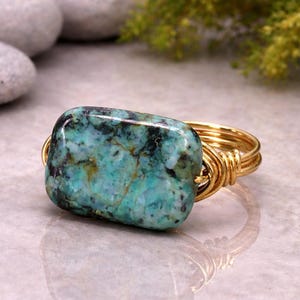 May include: A ring featuring a rectangular turquoise stone with dark matrix veining, secured by a gold-toned wire-wrapped band. The stone displays shades of teal, blue, and green, while the metal band consists of multiple coiled wire strands.