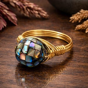 May include: A ring featuring a round, iridescent abalone shell mosaic centerpiece with small, square, multi-colored tiles. The band is constructed from multiple strands of gold-toned wire that are wrapped around the sides of the setting.