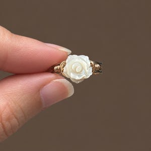 May include: A wire-wrapped ring featuring a carved white mother-of-pearl rose centrepiece. The band is constructed from thin gold-toned wire coiled around a darker metallic base, creating a layered appearance with a floral focal point.