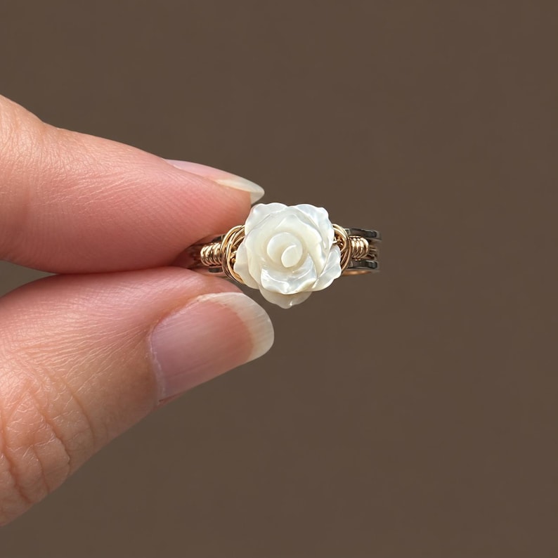 May include: A wire-wrapped ring featuring a carved white mother-of-pearl rose centrepiece. The band is constructed from thin gold-toned wire coiled around a darker metallic base, creating a layered appearance with a floral focal point.