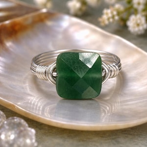 May include: A handcrafted ring featuring a faceted, square-cut, emerald-green gemstone. The stone is set in a silver-colored wire wrap design. The ring is displayed on a pearlescent surface, showcasing the jewelry's elegant design.