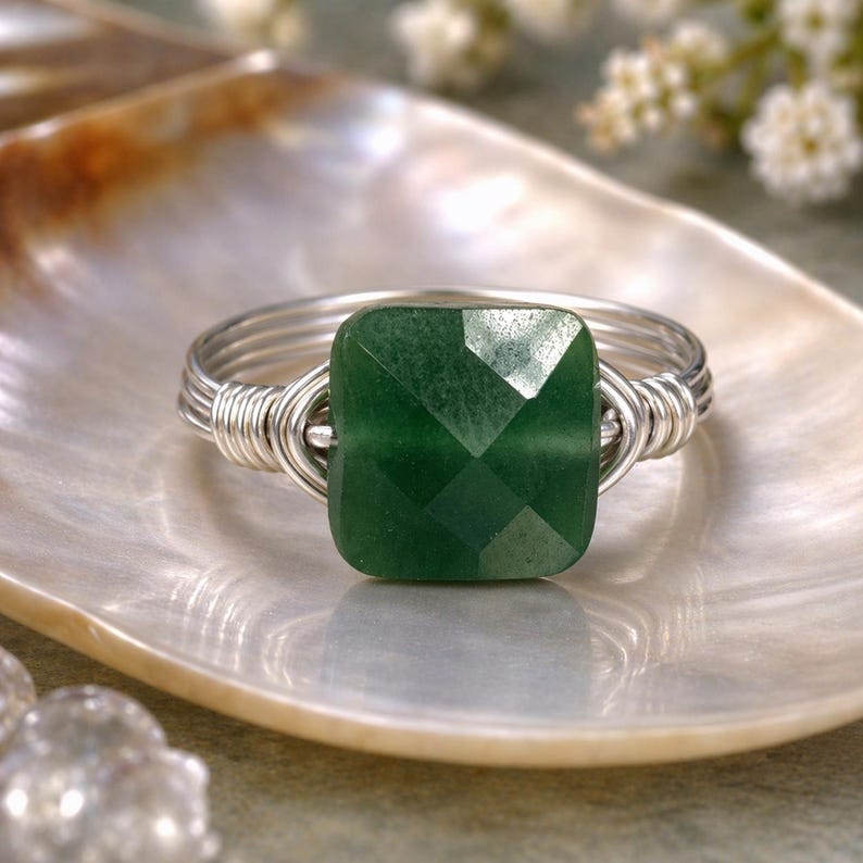 May include: A handcrafted ring featuring a faceted, square-cut, emerald-green gemstone. The stone is set in a silver-colored wire wrap design. The ring is displayed on a pearlescent surface, showcasing the jewelry's elegant design.