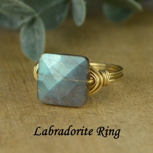May include: A gold-toned wire-wrapped ring featuring a square, faceted labradorite stone. The stone has a mix of blue, gray, and green hues. The text "Labradorite Ring" is displayed below the ring.