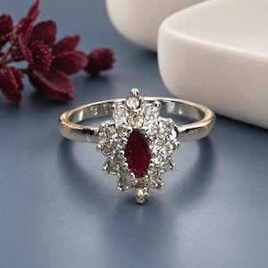 May include: A silver-toned ring featuring a central marquise-cut deep red gemstone surrounded by a cluster of small, clear, round-cut stones arranged in a pointed, elongated floral-inspired shape on a polished metal band.