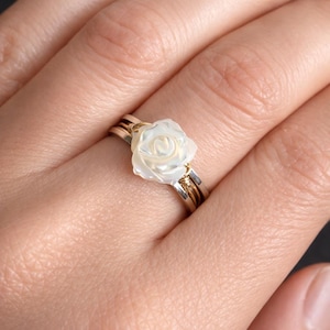 May include: A ring featuring a carved white mother-of-pearl rose centrepiece. The band is constructed from multiple thin, layered wires in alternating metallic tones of silver and gold, which are wrapped together to secure the floral element.
