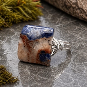May include: A handcrafted ring featuring a square-cut, polished stone with blue, brown, and cream hues. The stone is set in a silver-colored, wire-wrapped band. The ring is displayed on a reflective gray surface, showcasing the natural stone's unique patterns.