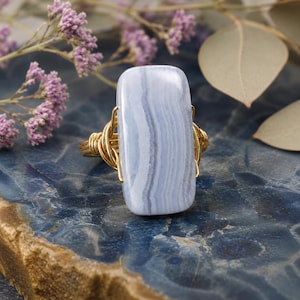 May include: A gold-toned wire-wrapped ring featuring a rectangular, light blue gemstone with subtle white and blue striations. The ring is set against a dark blue, textured surface, with sprigs of purple flowers and dried leaves in the background.