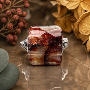 May include: A square-cut gemstone ring with a clear, reddish-brown stone. The stone is set in a silver wire wrap. The ring is displayed on a brown surface, with dried flowers and berries in the background. A unique piece of jewelry.