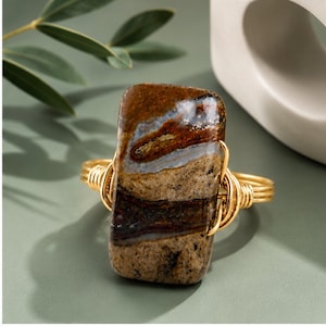 May include: A ring featuring a rectangular, polished brown and tan agate stone with visible banding and inclusions. The stone is secured by a gold-toned wire-wrapped band that coils around both sides of the gemstone, creating a textured metal finish.