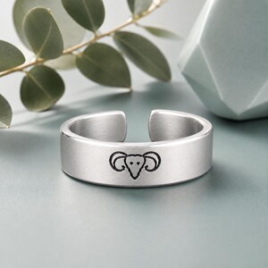 May include: A silver-toned metal ring with an open-ended band design. The outer surface of the ring features a centred, black-engraved graphic of a ram head with prominent curved horns, showcasing a smooth, matte finish across the band.