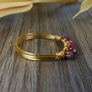 May include: A gold-toned wire-wrapped ring featuring three thin, parallel bands that converge at a focal point. This section is adorned with a cluster of small, faceted, deep magenta-colored gemstone beads secured by intricate gold wire coils.