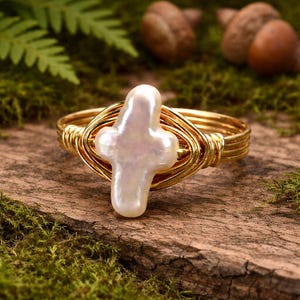 May include: A gold-toned wire-wrapped ring featuring a white, cross-shaped pearl. The ring's design includes multiple bands of gold wire, creating a textured and elegant look. The ring is set against a backdrop of moss and wood.