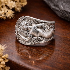 May include: A silver ring with a detailed relief of a reclining figure. The ring features a textured surface and intricate design, showcasing a classical or mythological theme. The ring is displayed on a dark wooden surface.