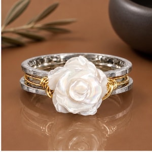 May include: A ring featuring a central white, iridescent, carved flower element. The band consists of two outer silver-toned hammered metal rings and two inner gold-toned wire-wrapped bands that secure the floral centrepiece in place.