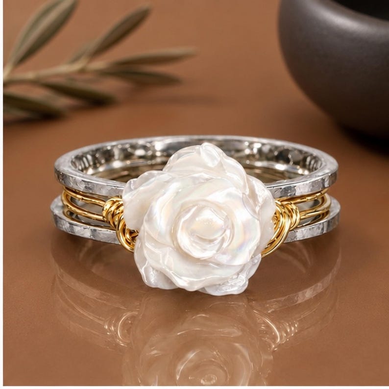 May include: A ring featuring a central white, iridescent, carved flower element. The band consists of two outer silver-toned hammered metal rings and two inner gold-toned wire-wrapped bands that secure the floral centrepiece in place.