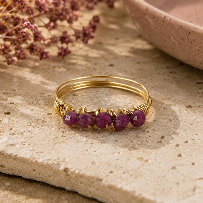 May include: A gold-toned wire-wrapped ring featuring five small, faceted, deep magenta-colored beads arranged in a row along the front. The band is constructed from multiple thin, coiled gold-colored wires that secure the beads in place.