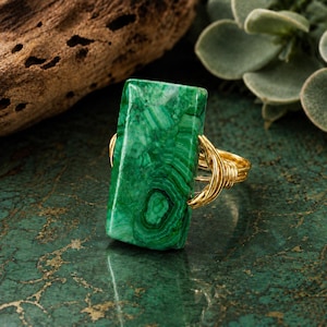 Pode incluir: Um anel retangular de malaquita com uma pedra verde polida que apresenta padrões naturais de faixas. A pedra é fixada por uma banda de fio dourado que se enrola em torno das laterais da gema para mantê-la firmemente no lugar.