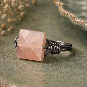 May include: A handcrafted ring featuring a square, faceted, peach-colored gemstone. The stone is set in a wire-wrapped, dark silver-toned band. The ring is displayed on a textured, green-toned surface, showcasing the jewelry's unique design.