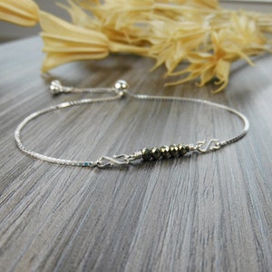 Peut inclure: Un bracelet en chaîne argent avec un fermoir coulissant et une rangée de perles de pyrite au centre.