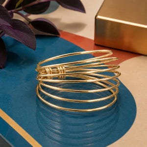 May include: A gold-toned multi-strand bangle bracelet featuring several thin, circular wire bands that overlap to create a layered appearance. The individual wire strands are secured together at one side by two small, tightly coiled gold wire accents.
