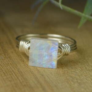 A silver ring with a square-shaped rainbow moonstone stone in the center, resting on a wooden surface.