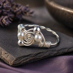 May include: A silver wire-wrapped ring featuring a spiral design with three faux pearls and two silver beads. The ring sits on a dark gray stone surface, with a blurred purple fabric in the background.