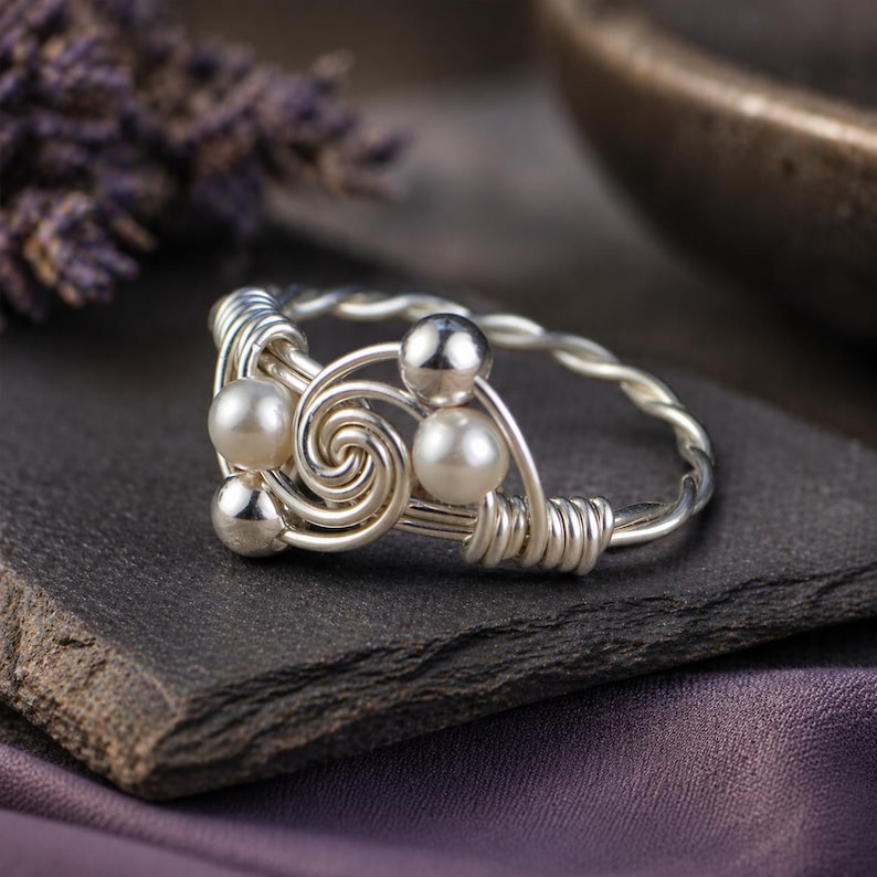 May include: A silver wire-wrapped ring featuring a spiral design with three faux pearls and two silver beads. The ring sits on a dark gray stone surface, with a blurred purple fabric in the background.