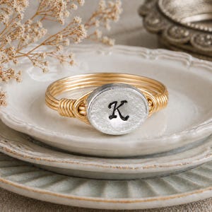 May include: A gold-toned wire-wrapped ring featuring a central oval-shaped silver-colored metal charm. The charm has a textured, matte finish and displays the letter K in a black, cursive script font centered on its surface.