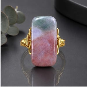 May include: A ring featuring a rectangular, polished stone with mottled pink, lavender, and green hues. The stone is secured by a gold-toned wire-wrapped setting that forms a decorative, coiled band around the sides of the central gemstone.