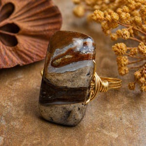 May include: A handcrafted ring featuring a rectangular stone with brown, white, and gray layers. The stone is wrapped in gold-colored wire, creating a unique and earthy jewelry piece. The ring is a statement piece, perfect for adding a touch of natural beauty to any outfit.