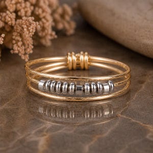 May include: A gold-toned ring with three bands and a series of silver-colored beads. The ring has a simple, elegant design, with the beads adding a touch of texture and visual interest. The ring is likely made of metal and is suitable for everyday wear.