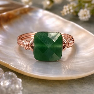 May include: A copper wire ring with a square, faceted, emerald-green gemstone. The ring is set on a pearlescent, shell-shaped dish, with soft focus flowers in the background. The ring's wire is wrapped around the gemstone.