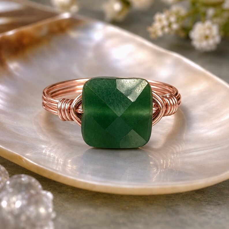 May include: A copper wire ring with a square, faceted, emerald-green gemstone. The ring is set on a pearlescent, shell-shaped dish, with soft focus flowers in the background. The ring's wire is wrapped around the gemstone.