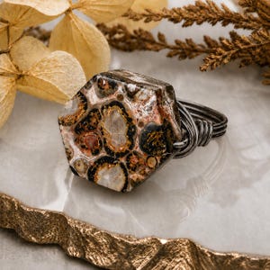 May include: A ring featuring a hexagonal, polished stone with a mottled pattern of black, brown, tan, and cream spots. The stone is secured by dark, oxidized metal wire wrapped around the sides to form a band, creating a textured, industrial look.