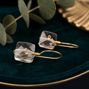 May include: A pair of clear, square-cut crystal earrings with gold-colored wire detailing. The earrings are displayed on a dark green surface, with a blurred background of greenery and a gold-colored decorative element.